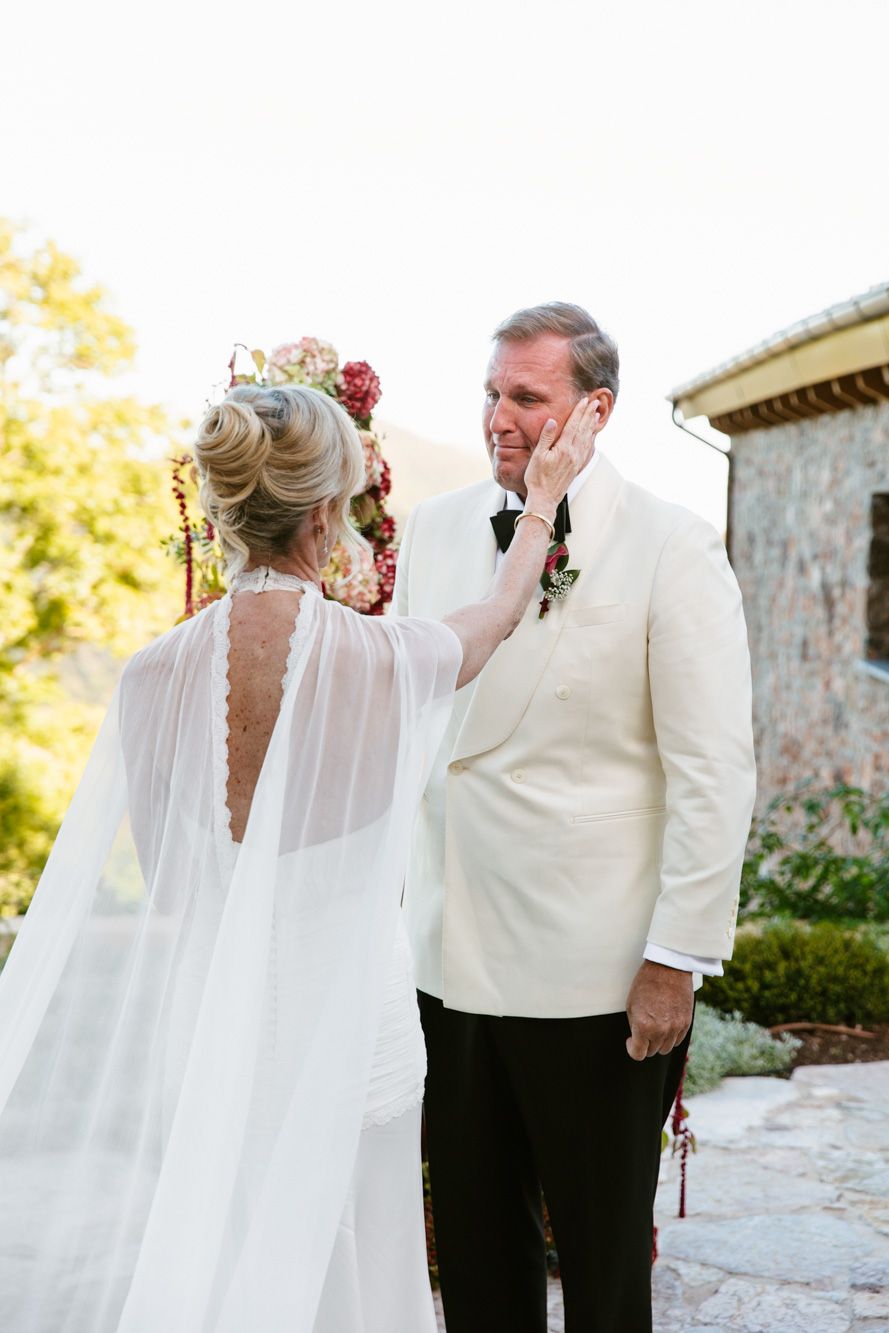 A bride in a sheer caped veil gently touches the cheek of an emotional groom in a white dinner jacket during a first look outdoors.