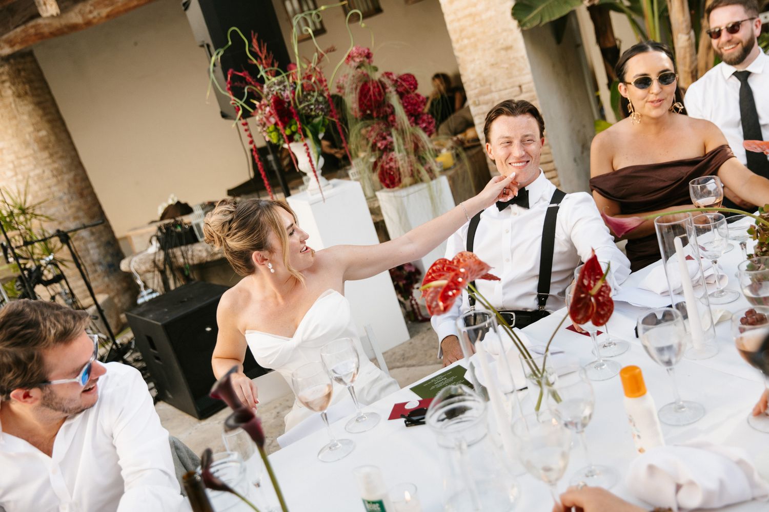 Bride and groom laughing at wedding reception dinner table with guests in elegant outdoor venue.