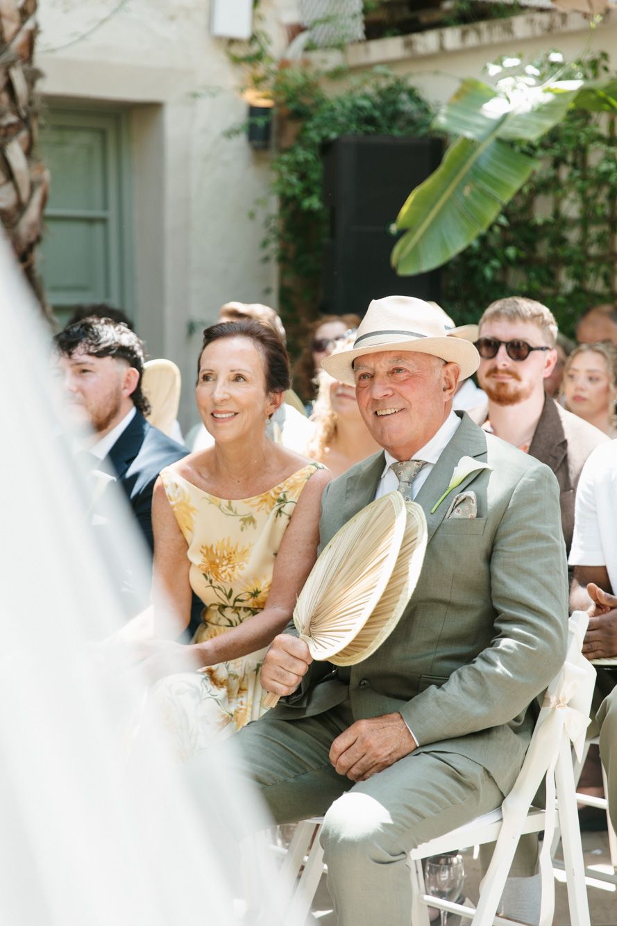 Wedding guests seated outdoors, older couple smiling, man in gray suit holding a palm leaf fan at a garden ceremony.