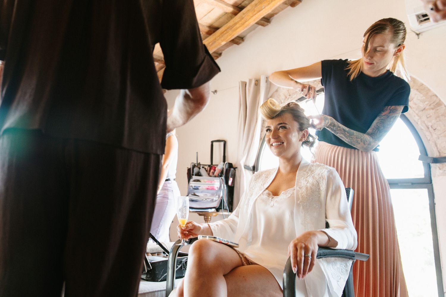 Bride smiling in white robe getting hair styled by hairdresser before wedding ceremony indoors.