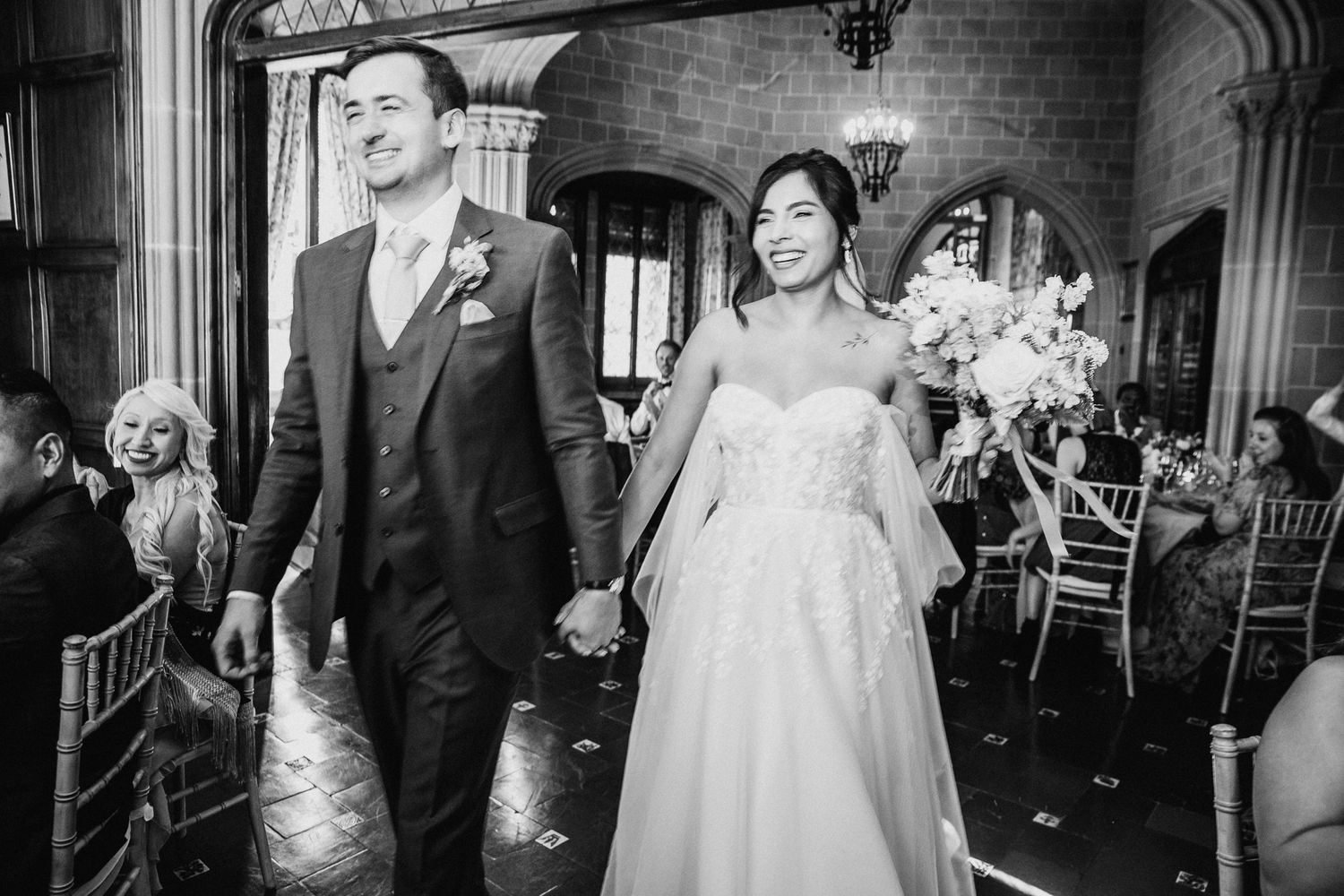 Black and white photo of a smiling bride and groom holding hands walking down the reception area at La Baronia