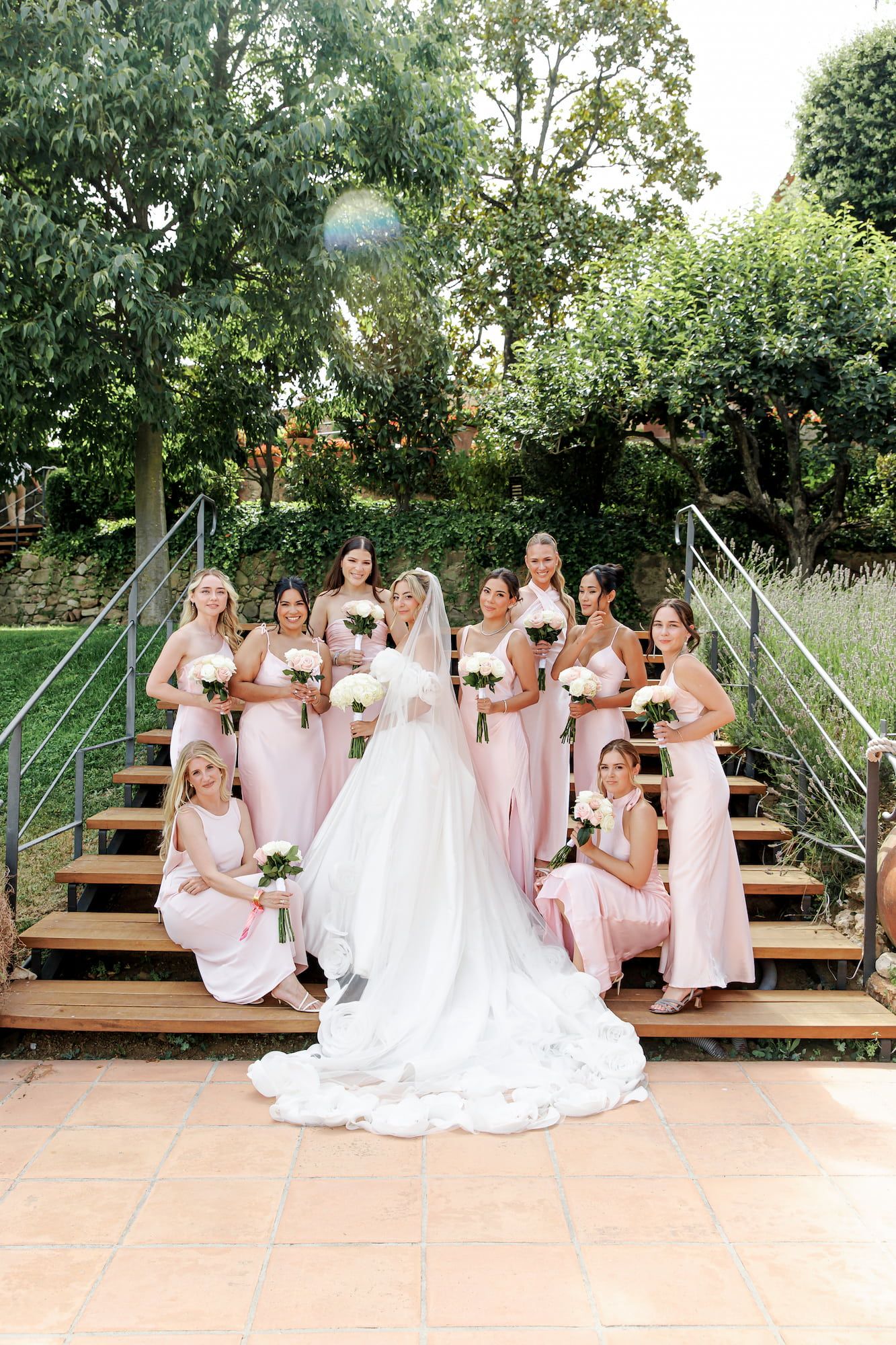 Bride in white gown poses with bridesmaids in pink dresses on outdoor steps surrounded by lush greenery.
