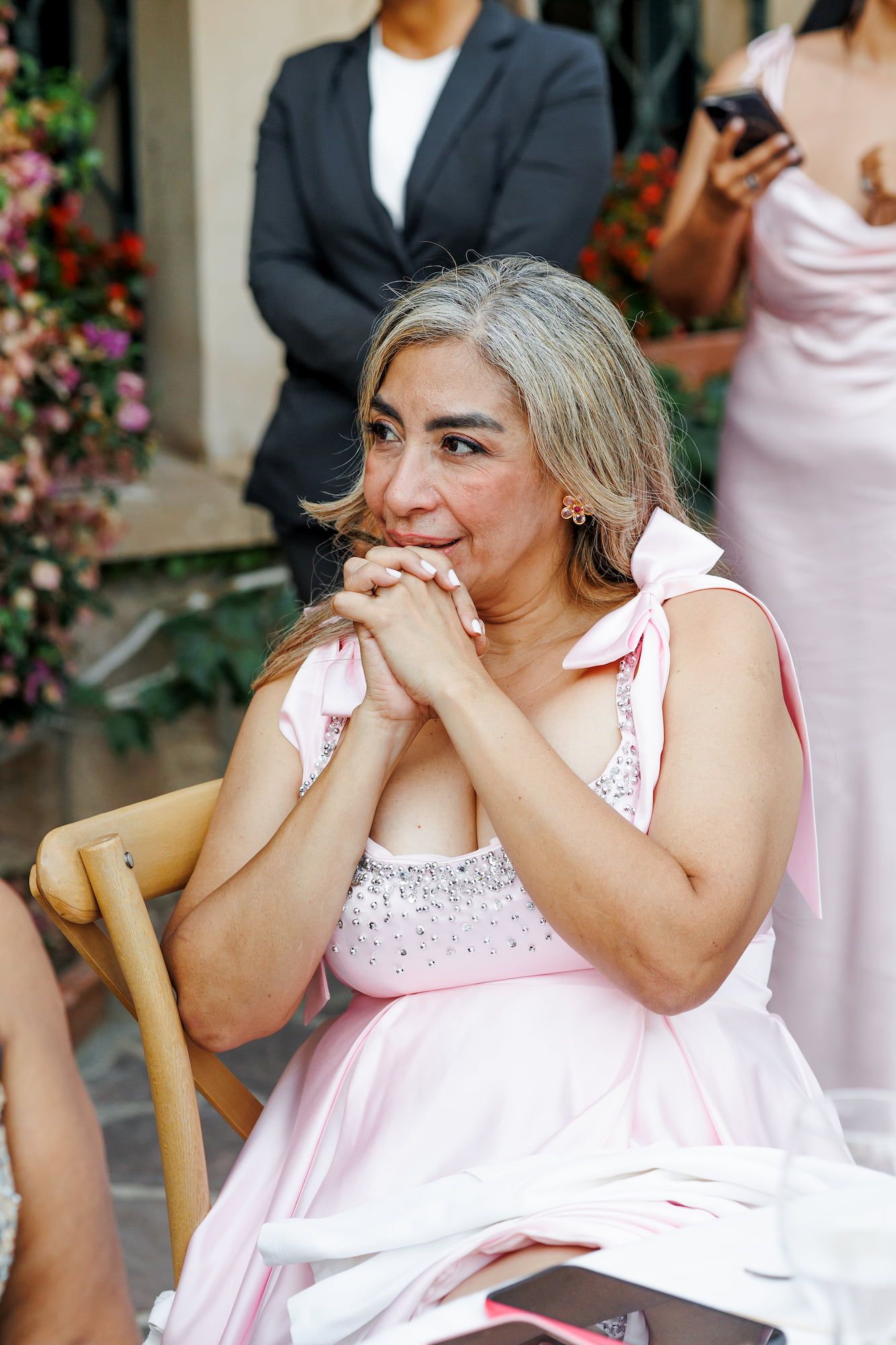 Emotional woman in pink dress with bow detail sits clasping hands at a wedding ceremony.