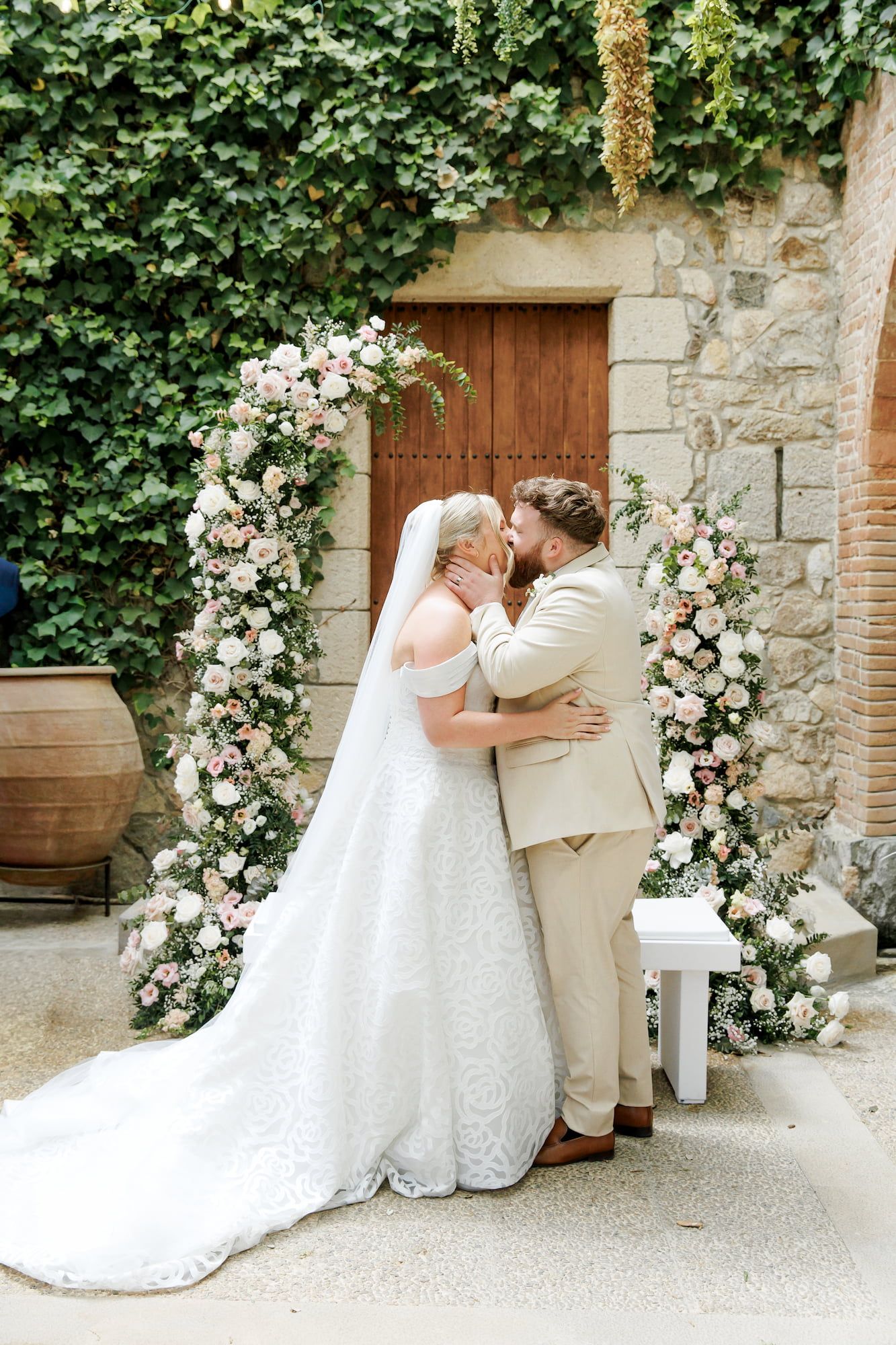 Bride and groom share first kiss under floral arch with pink roses at outdoor wedding ceremony.