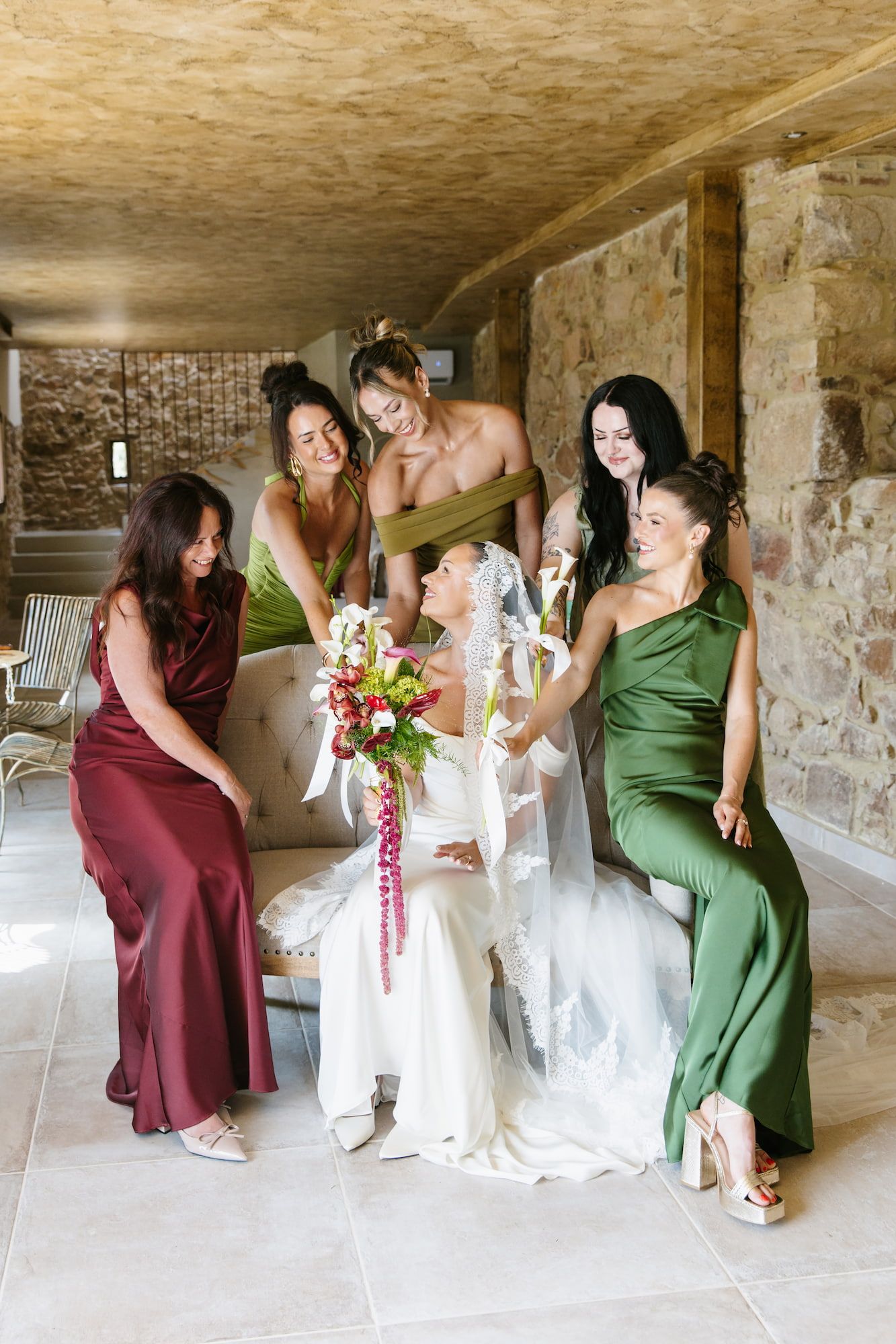 Bride seated with bouquet surrounded by bridesmaids in green and burgundy satin gowns in a rustic stone venue.