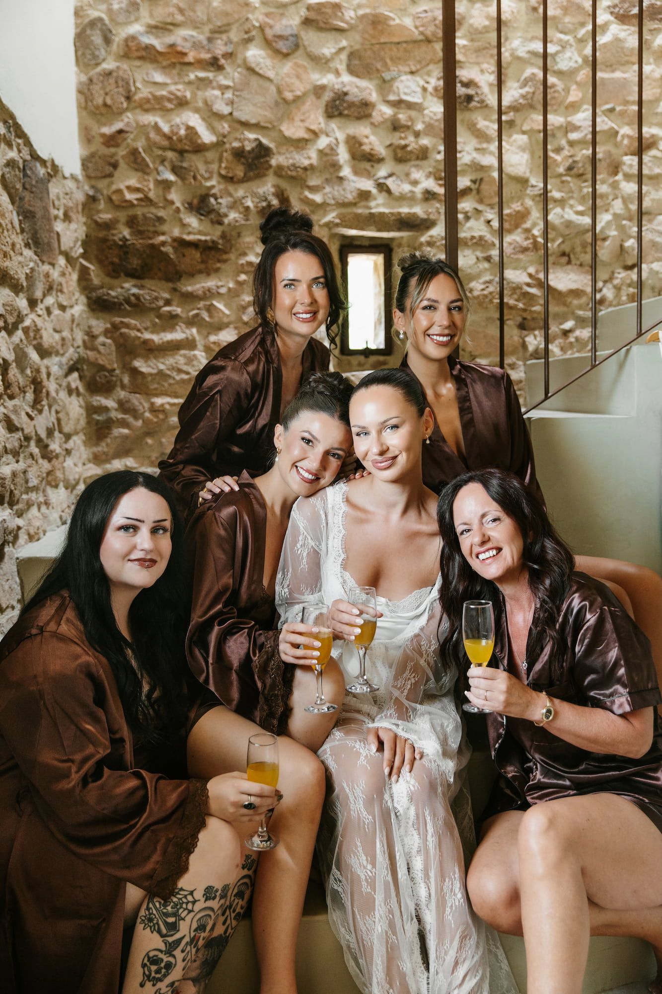 Bride in white robe with bridesmaids in brown satin robes holding champagne glasses, posing in a stone-walled venue.