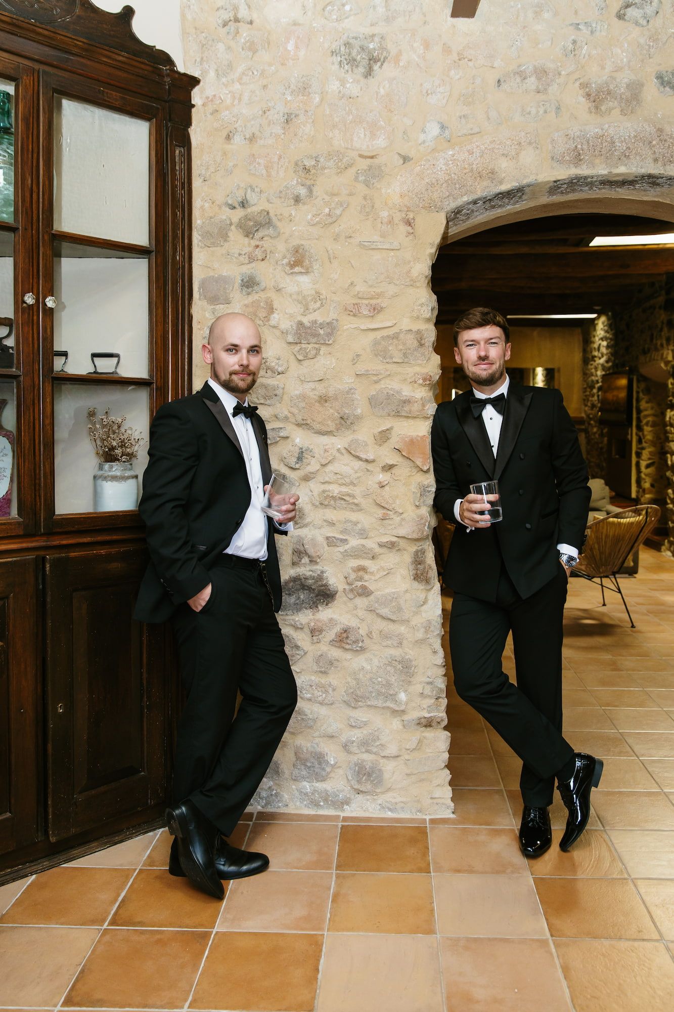 Two men in black tuxedos with bow ties holding drinks, posing against a rustic stone wall in an elegant venue.
