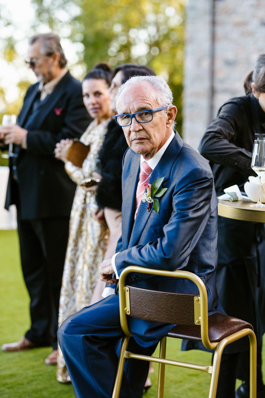 Elderly man in glasses and a blue suit with a floral boutonniere sits on a gold chair at an outdoor wedding as guests stand behind him.
