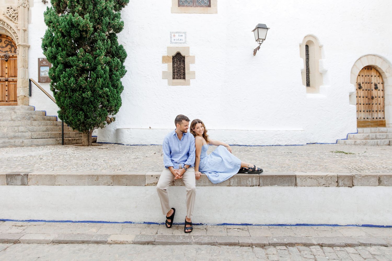 A romantic couple sits together on the white stone steps of the Mediterranean church courtyard with cypress trees.