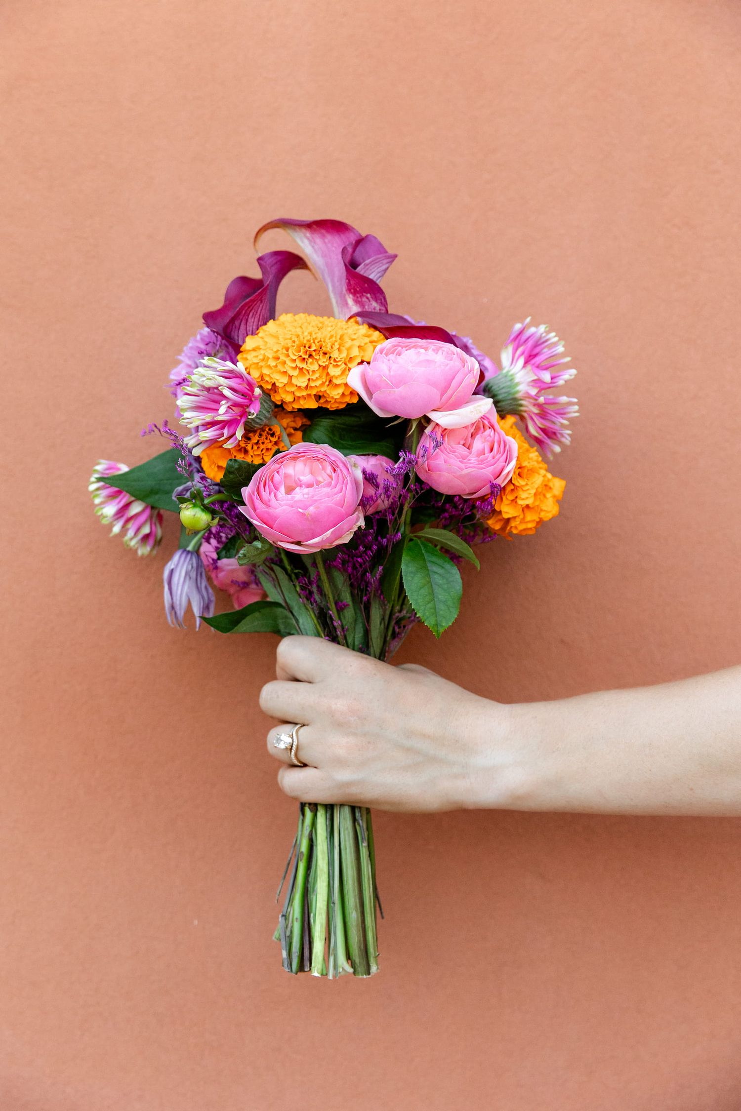 A hand holds a vibrant bouquet of pink ranunculus, purple daisies, and orange marigolds against a peach-colored background.