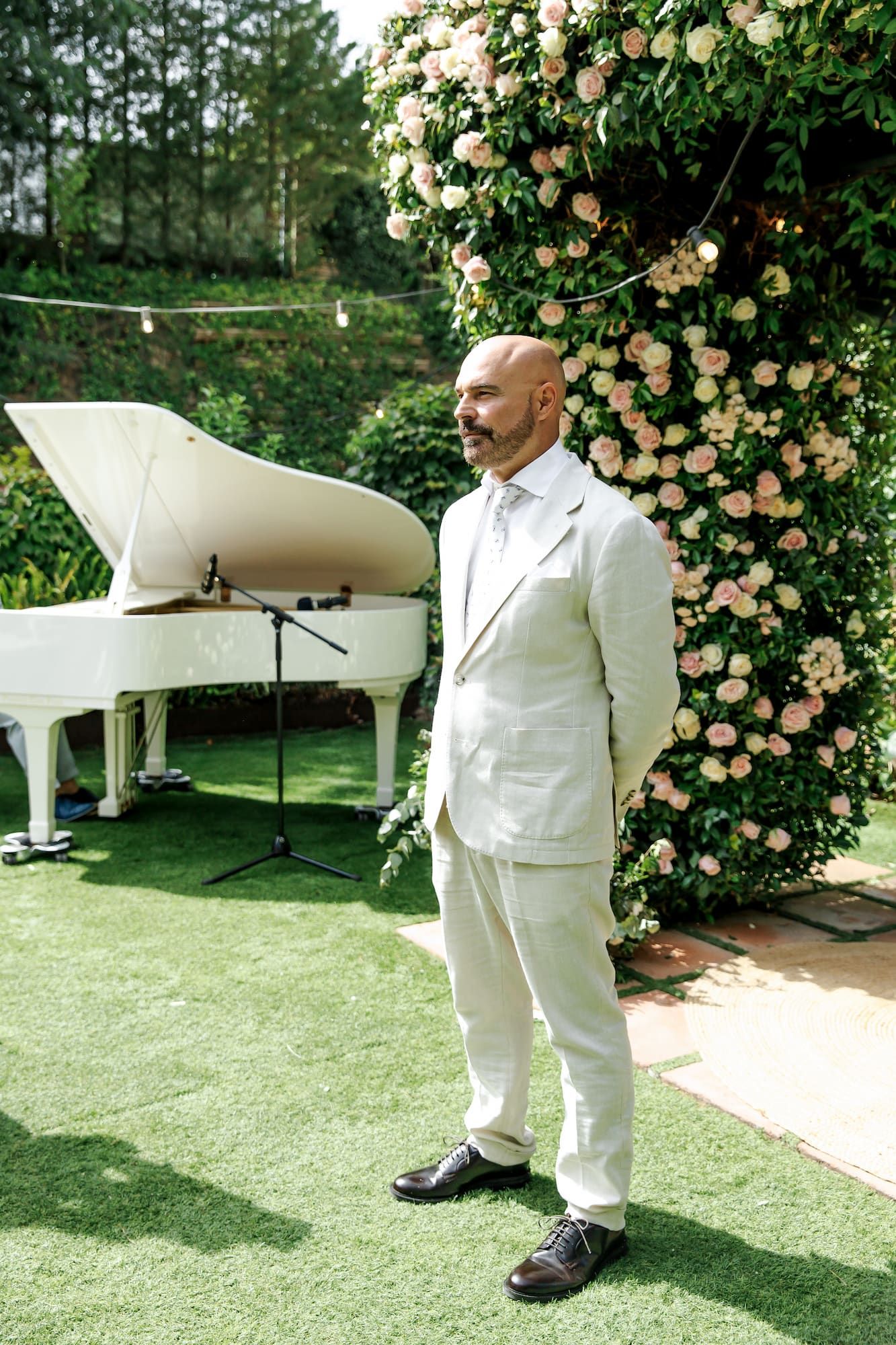 Elegant man in white suit stands outdoors near a white grand piano and floral wall at a wedding event.