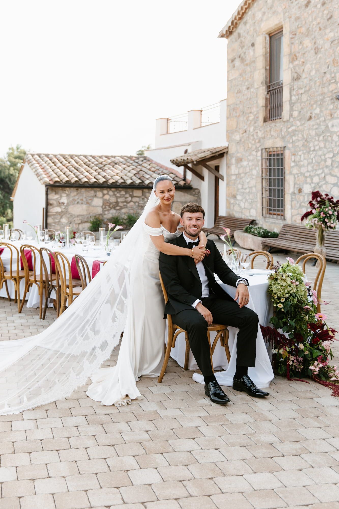 Bride in white gown and veil poses with groom in black suit at elegant outdoor wedding reception venue.