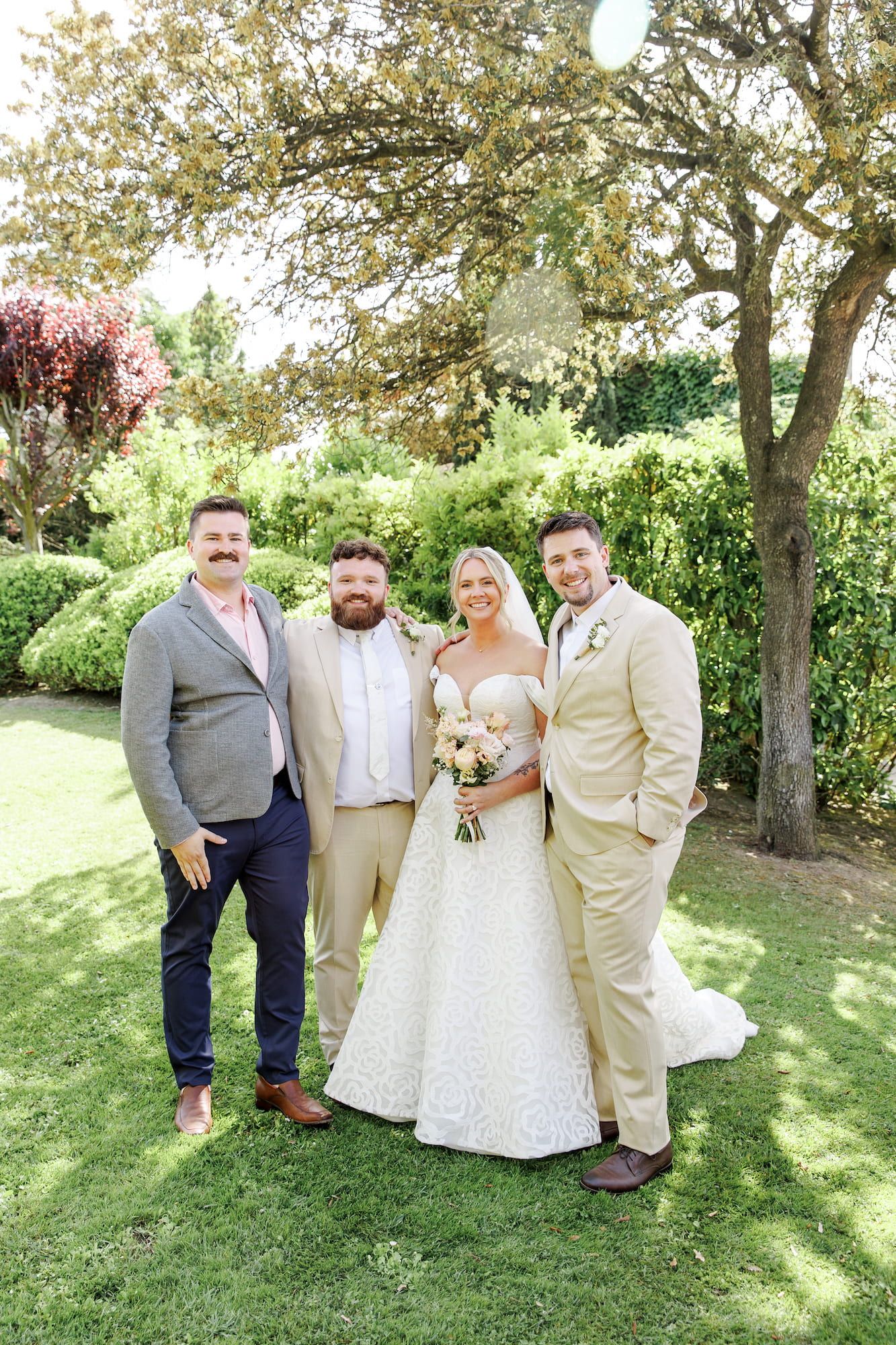 Bride in white gown with groom and two guests posing in a lush garden on a sunny wedding day.