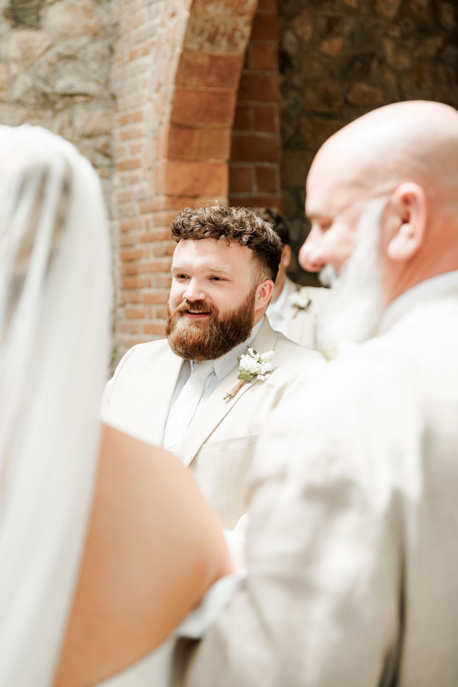 Groom in white suit smiling at bride during outdoor wedding ceremony near rustic brick archway.