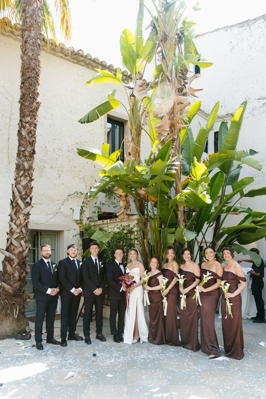 Wedding party poses outdoors at Masia Victoria with groomsmen in black suits and bridesmaids in brown gowns