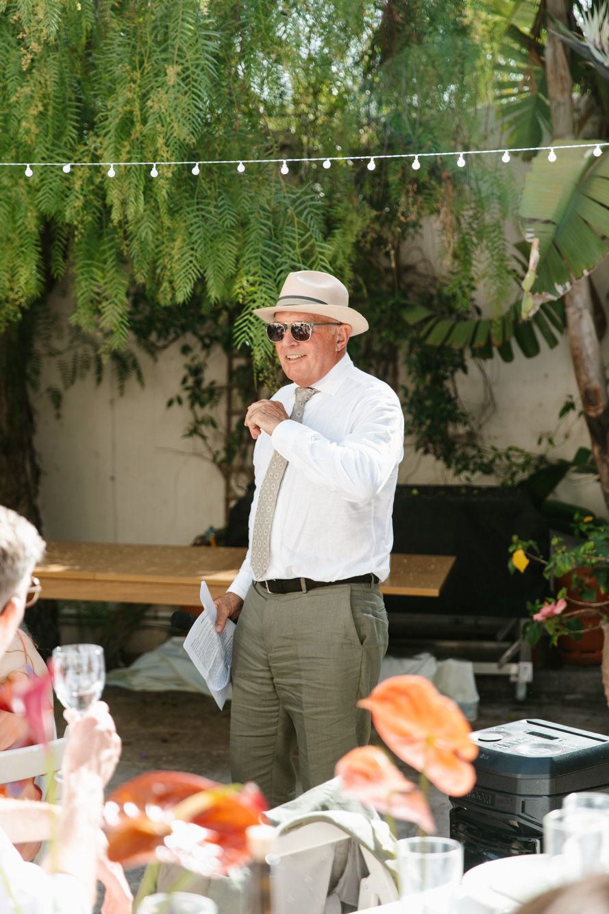 Elderly man in white shirt, tan hat, and sunglasses speaking at an outdoor garden wedding reception.