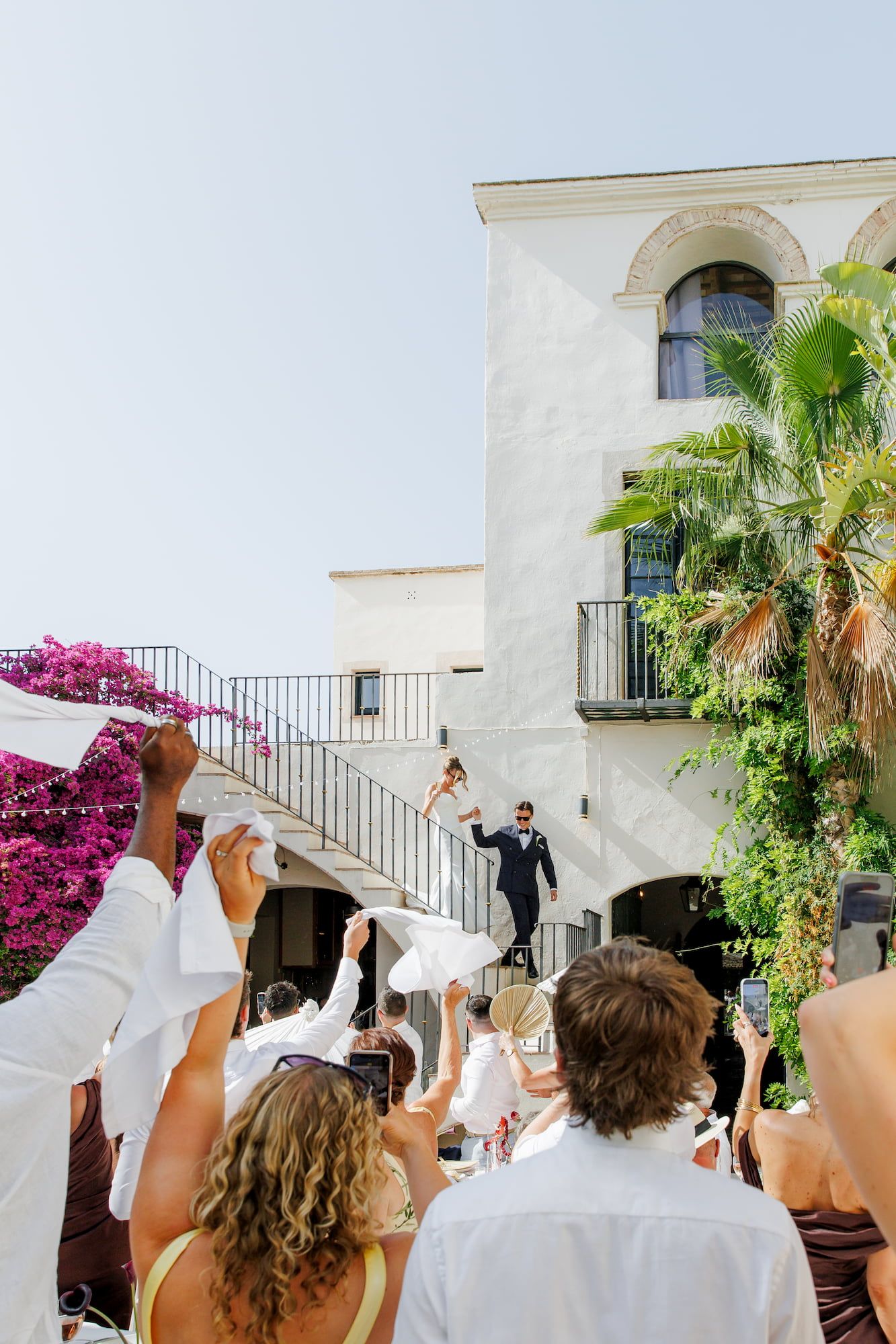 Wedding guests wave white napkins as newlyweds descend the staircase at Masia Victoria near Barcelona.