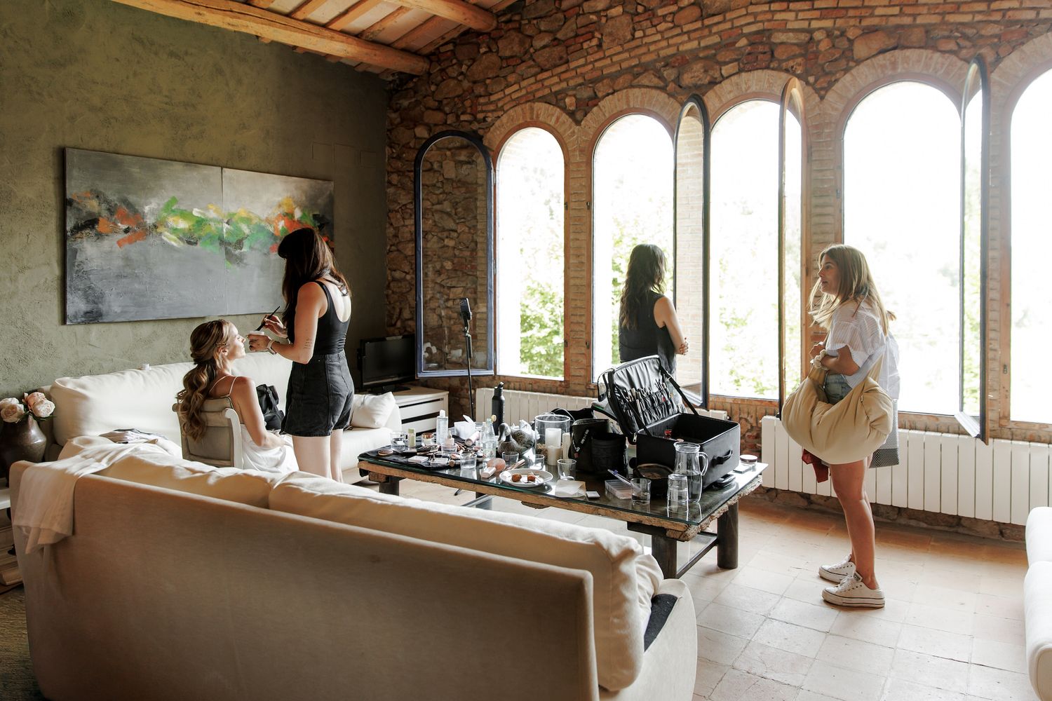 Women gather in a rustic chic living room with arched windows, brick walls, and modern art, enjoying a social gathering.