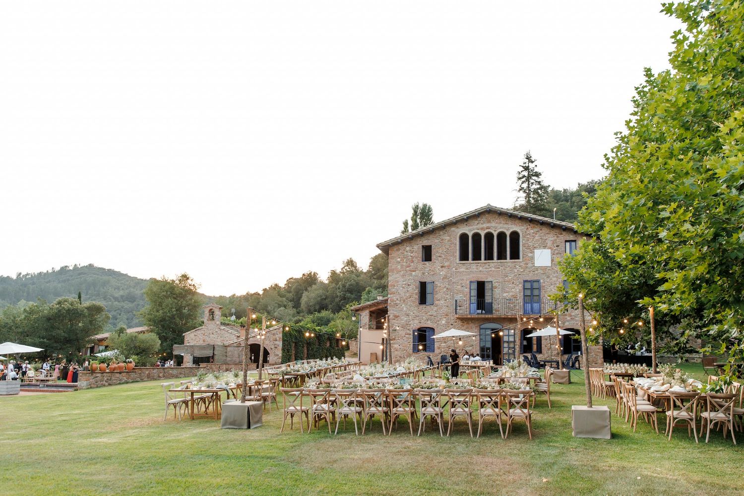 Outdoor wedding reception setup on green lawn beside rustic stone villa surrounded by trees and rolling hills.