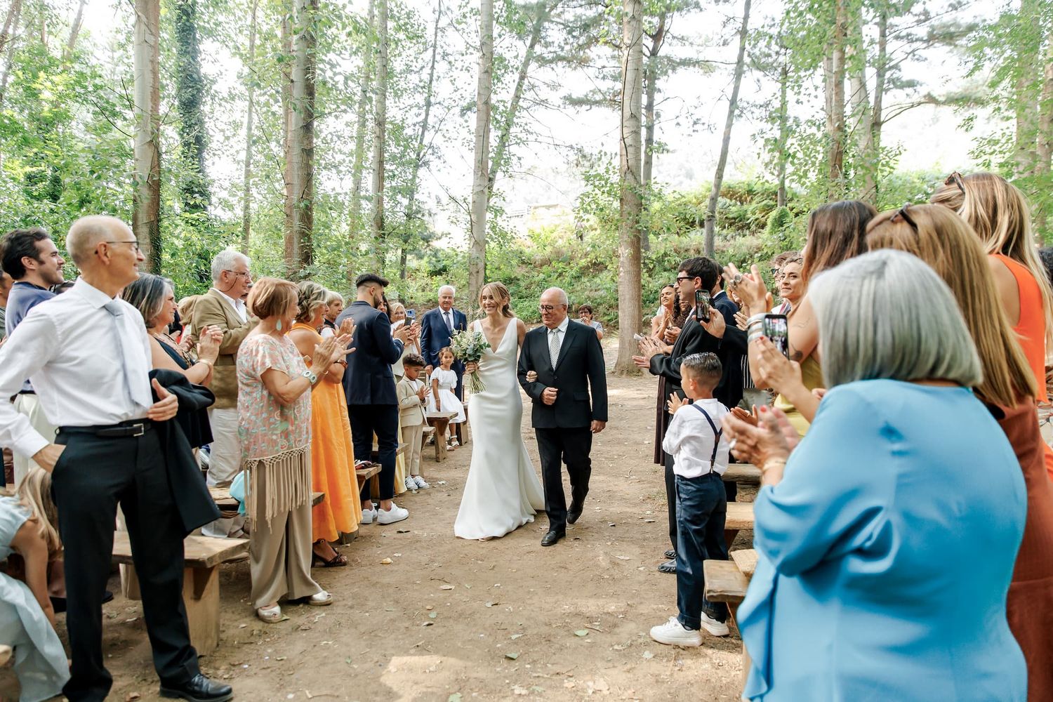 Bride walks down outdoor forest aisle with guests applauding at a woodland wedding ceremony.