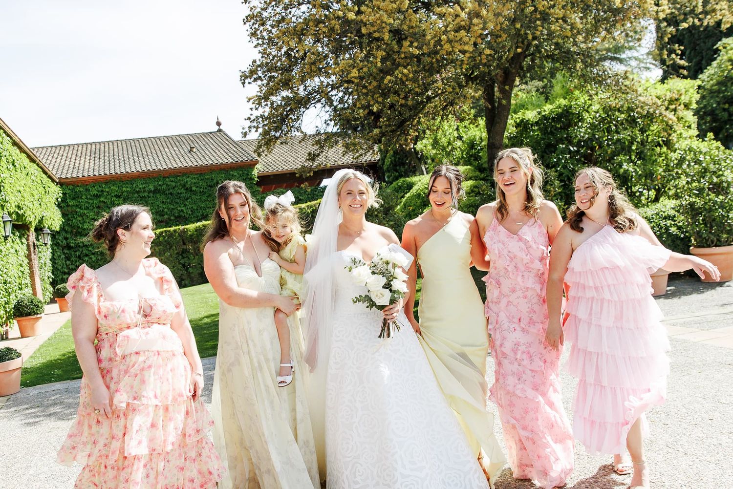 Bride in white gown poses with six bridesmaids in pink and cream dresses outdoors at a sunny wedding venue.