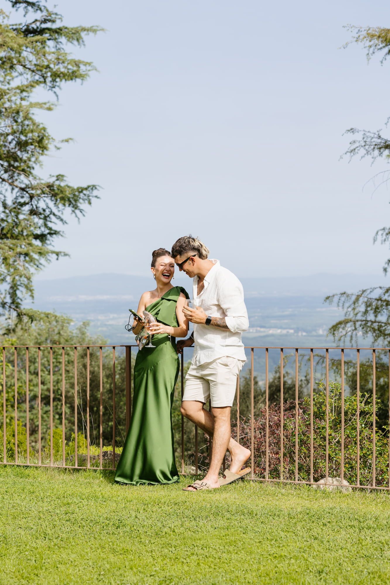 Couple in green dress and white outfit enjoying drinks on a scenic garden terrace with panoramic views.