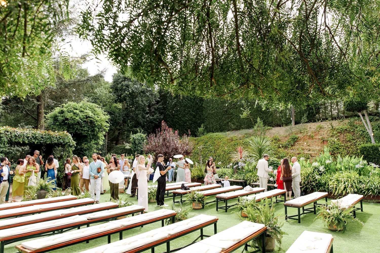 Outdoor wedding ceremony with wooden benches on green lawn surrounded by lush tropical garden trees and guests.