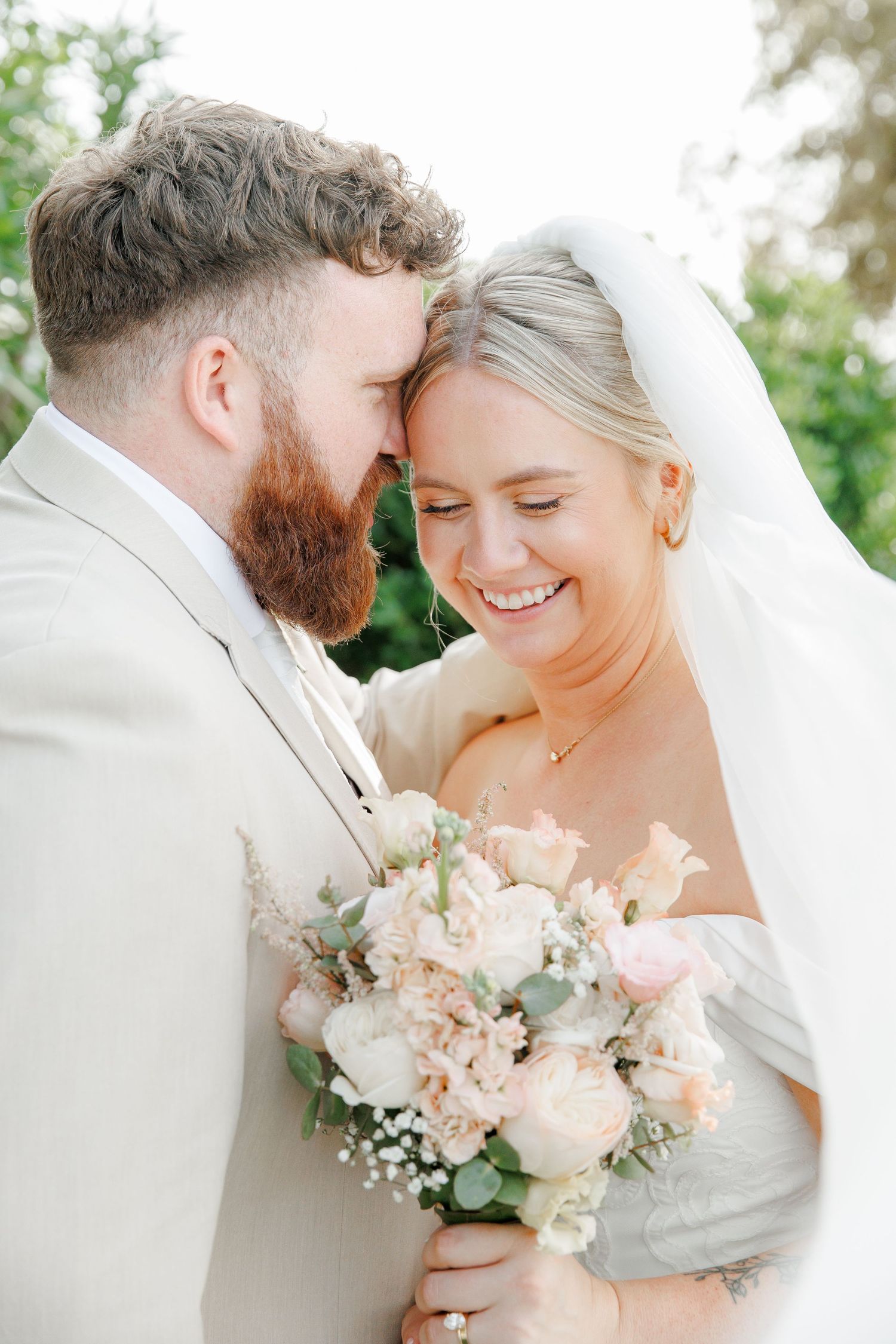 A smiling bride holding a blush bouquet leans forehead-to-forehead with a bearded groom in a cream suit outdoors.