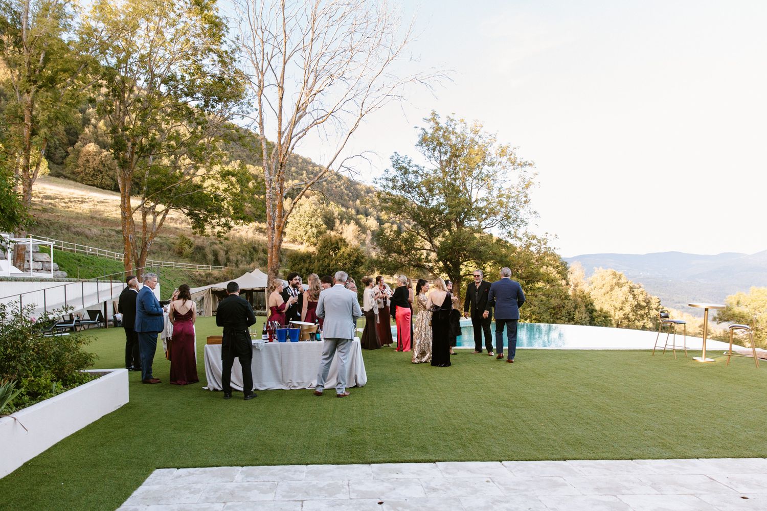 Wedding guests gather around a white-draped table on a green lawn beside an infinity pool with scenic mountain and valley views.
