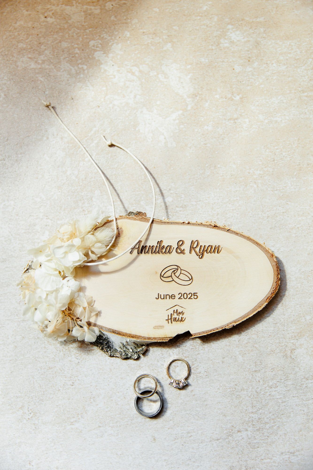 A rustic wood slice wedding favor with engraved names and rings lies beside delicate white flowers and engagement rings.