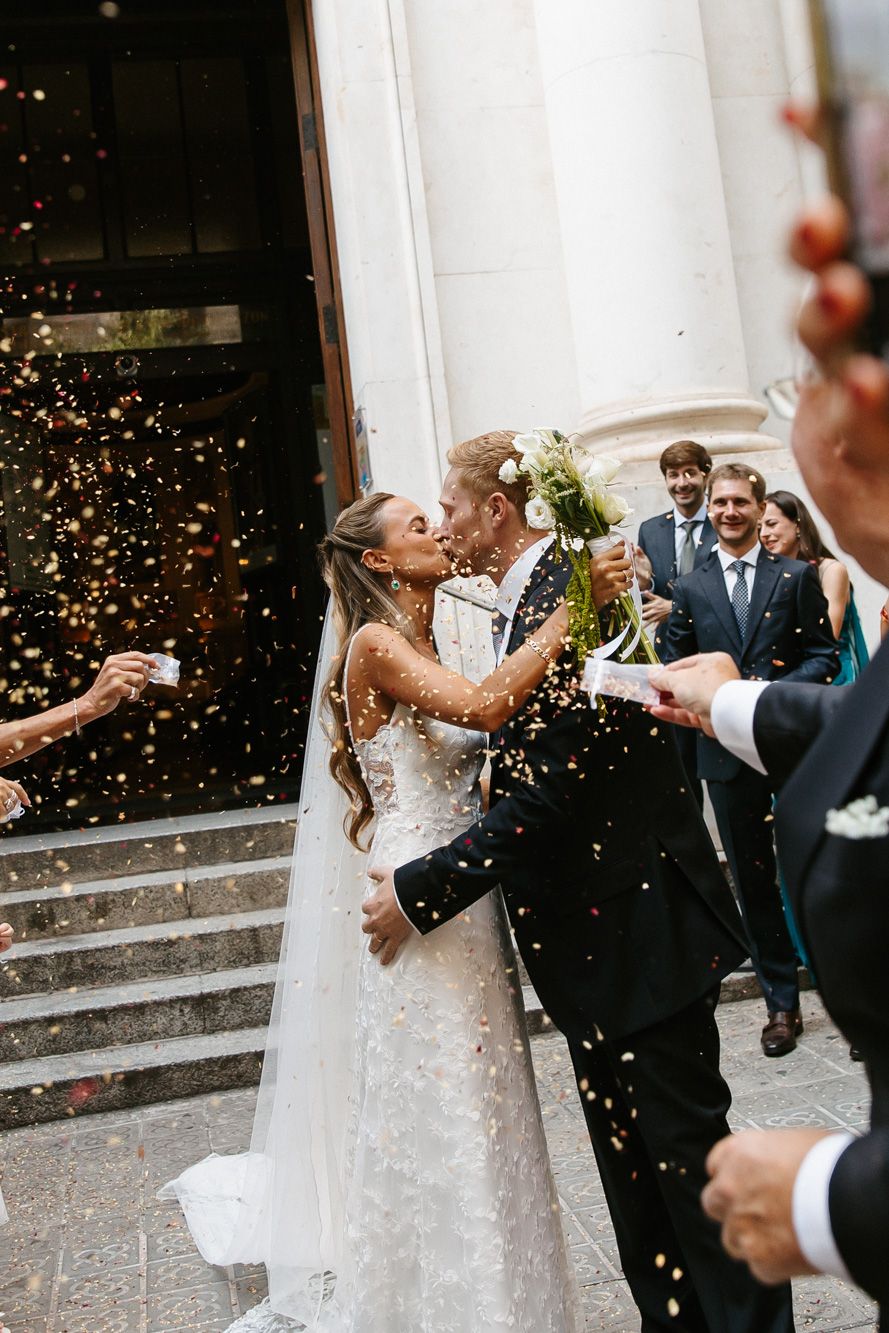 Bride and groom share a kiss outside church as guests toss golden confetti during joyful wedding ceremony exit.