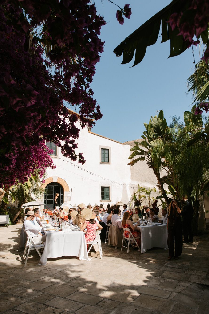 Outdoor wedding reception at a Mediterranean villa with guests dining at white-clothed tables, framed by bougainvillea.