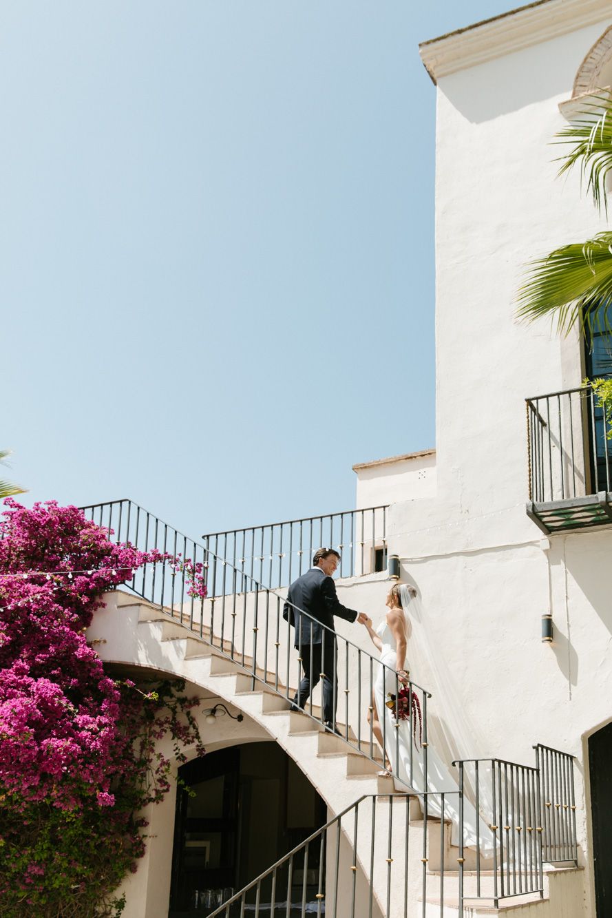 Bride and groom walking up elegant spiral staircase with pink bougainvillea at Masia Victoria