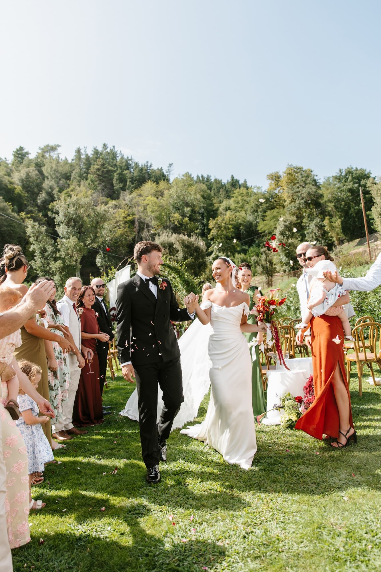 Newlyweds walk down outdoor aisle as guests celebrate with confetti at sunny garden wedding ceremony.