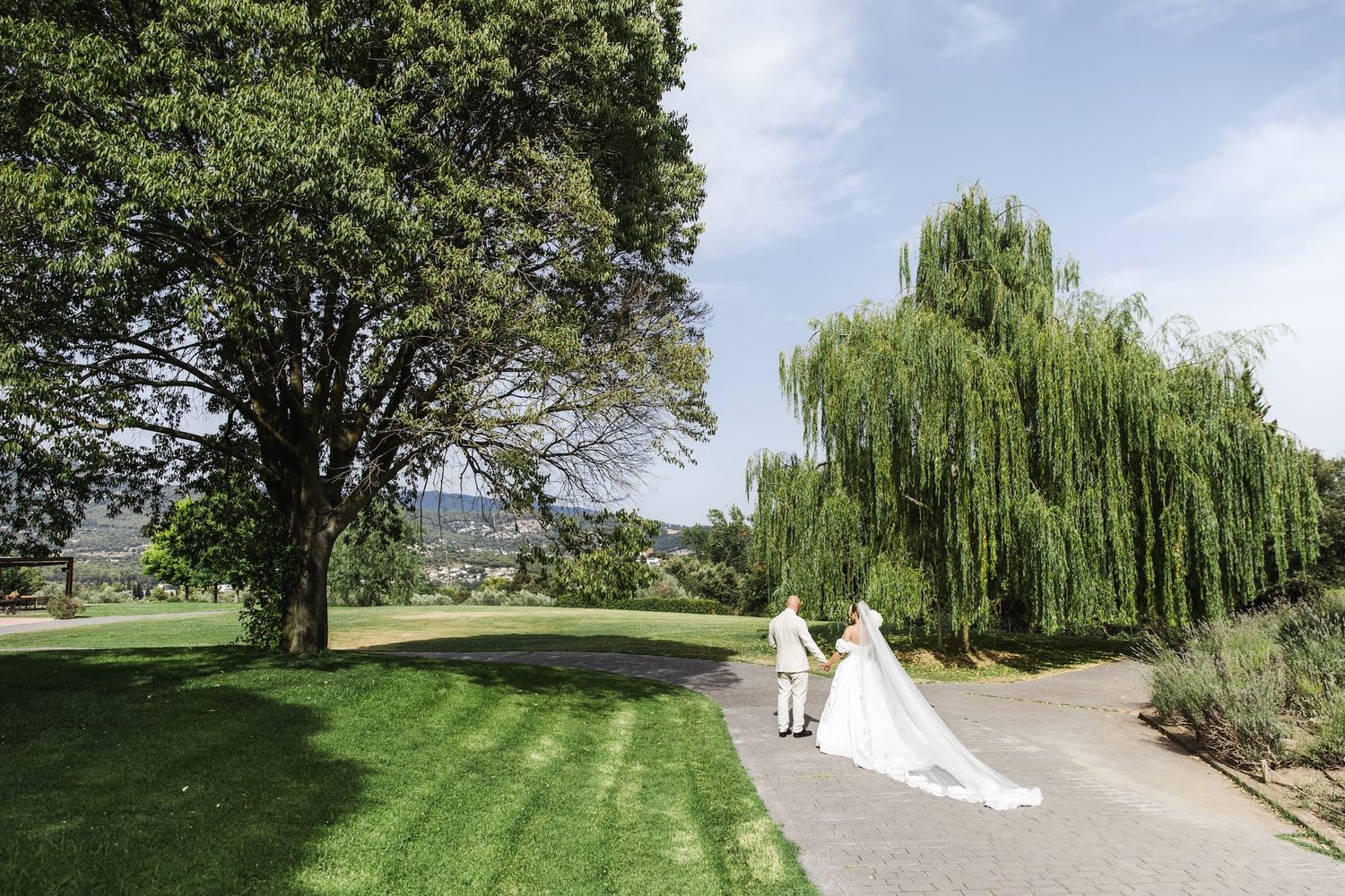 Bride and groom walking hand in hand along a garden path surrounded by lush green trees on a sunny wedding day.