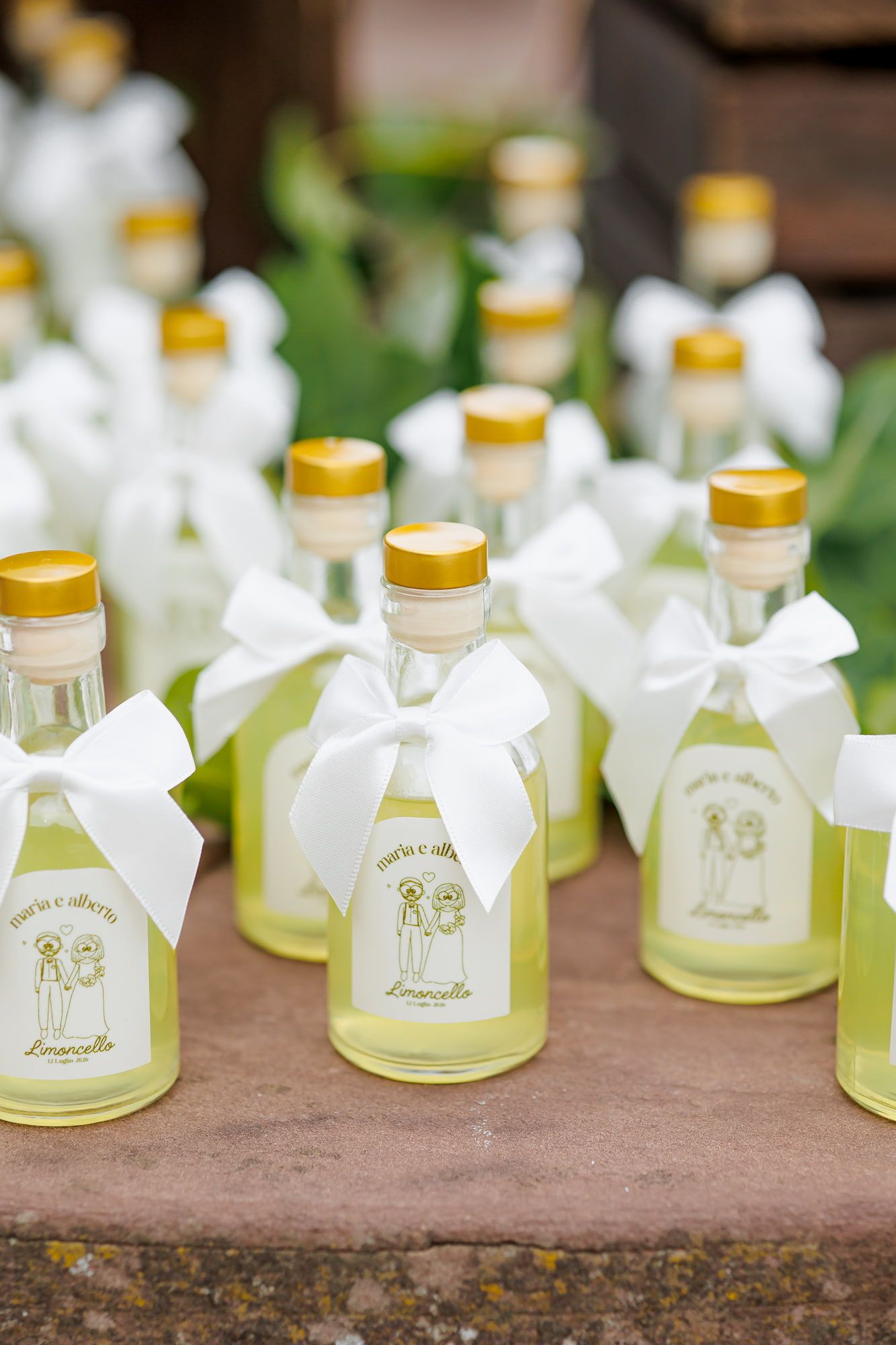Mini limoncello bottles with white ribbon bows and custom labels displayed as wedding favors on a stone surface.