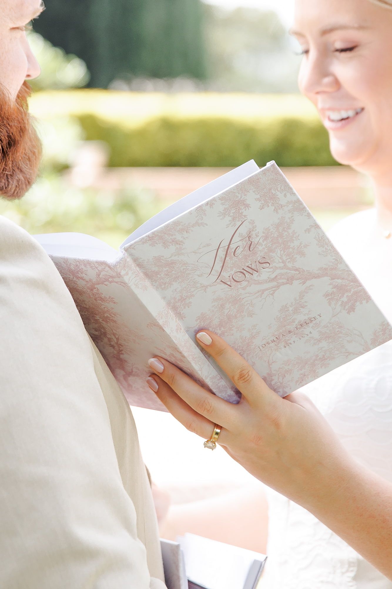 Smiling bride reads wedding vows from an elegant floral-covered booklet beside her bearded groom outdoors.