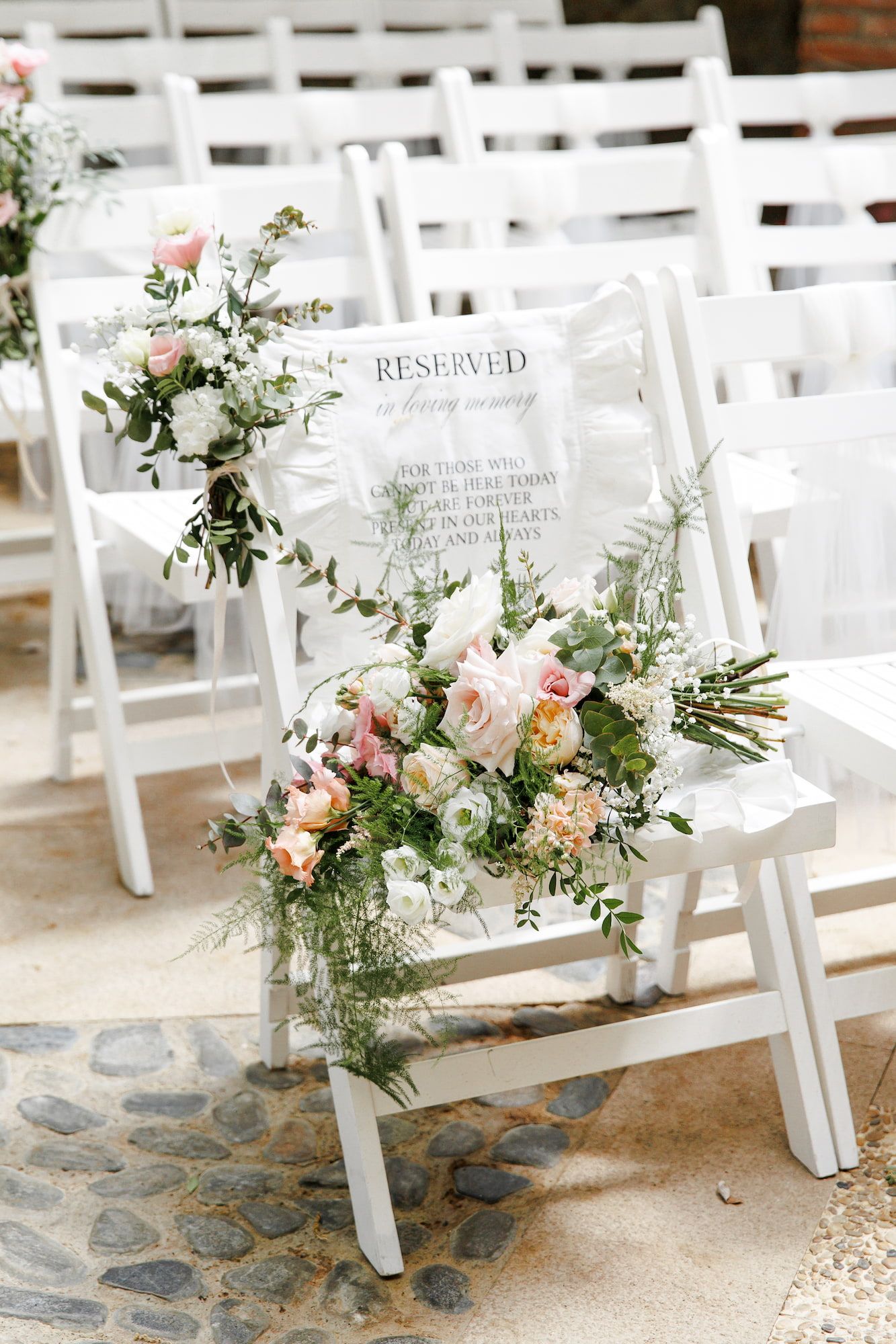 White folding chairs with floral arrangements and a reserved sign at an outdoor wedding ceremony.