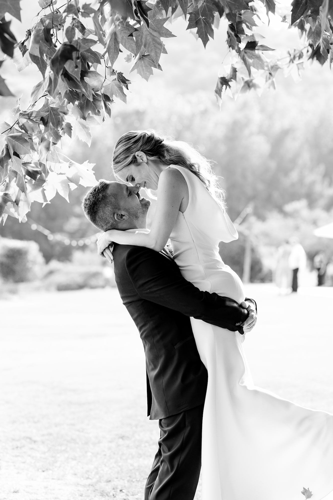 Black and white photo of groom lifting bride under leafy tree in garden wedding portrait.