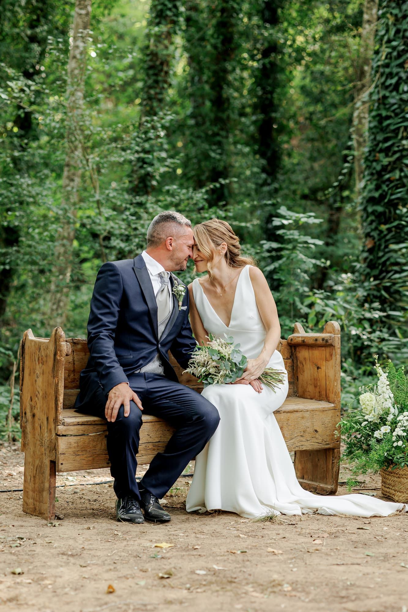 A bride and groom share a romantic moment on a rustic wooden bench surrounded by lush greenery on their wedding day.