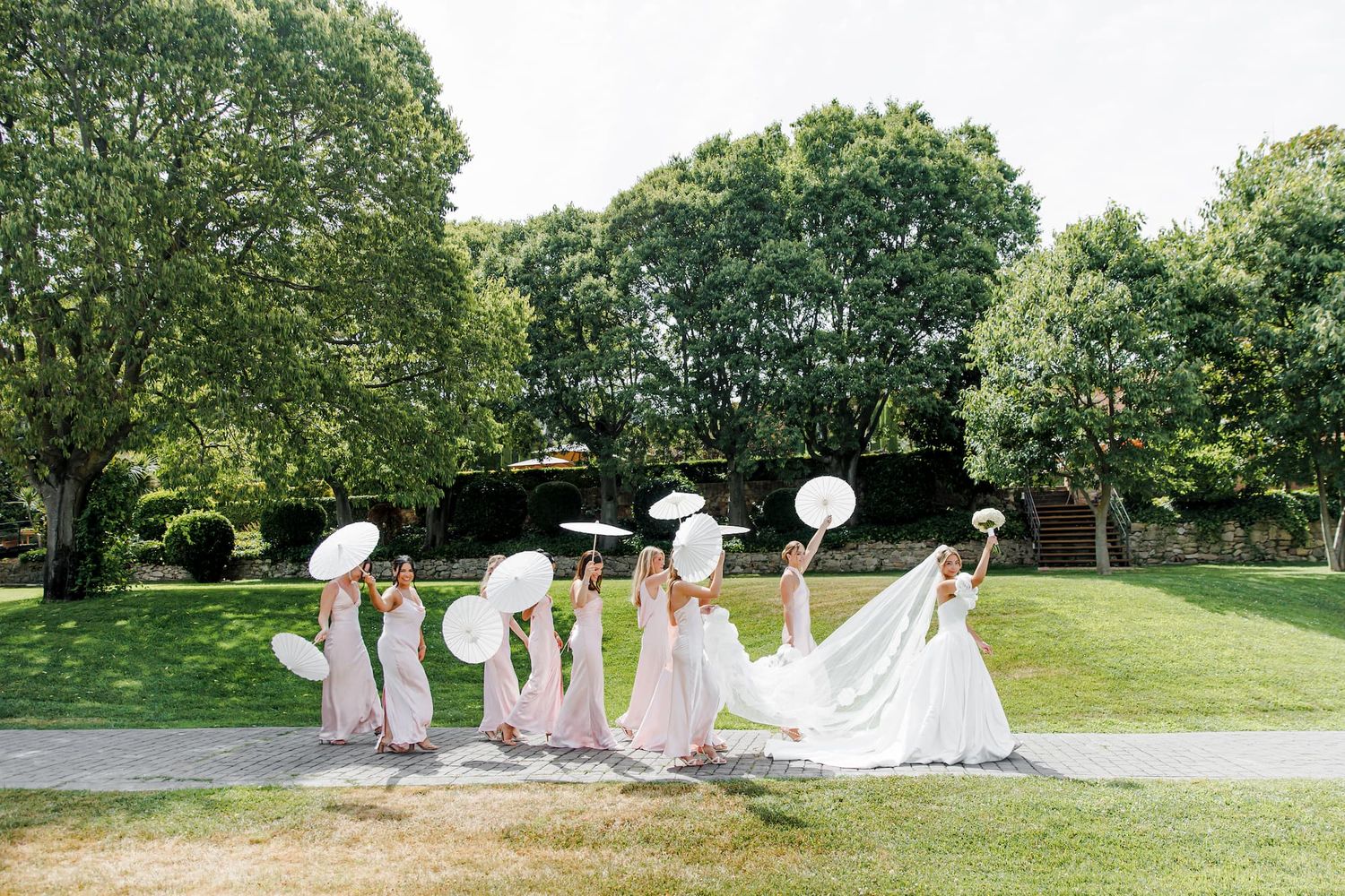 Bride and bridesmaids in pink dresses holding white parasols walking on green lawn at outdoor wedding.