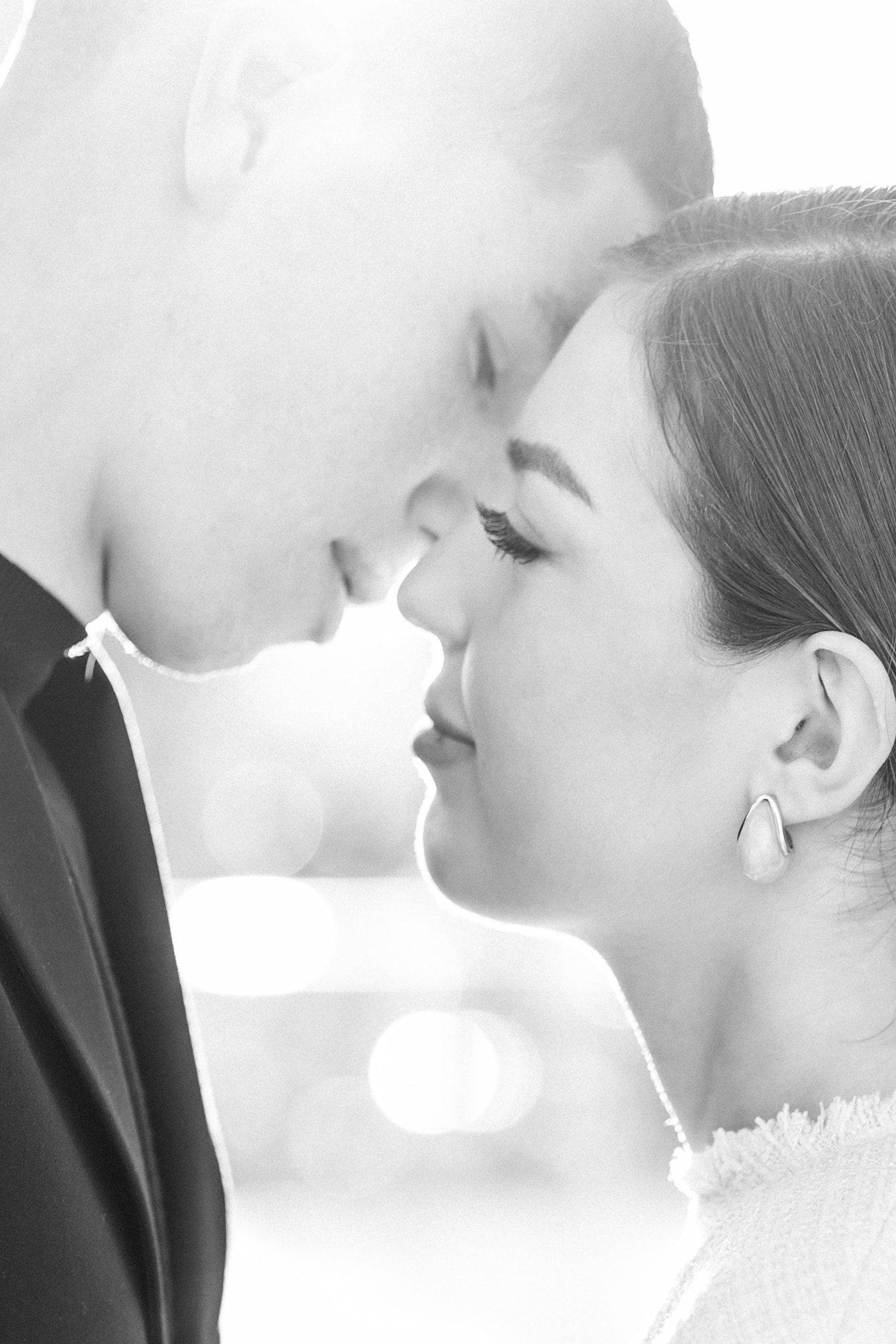 Black and white photo of a romantic couple facing each other with foreheads almost touching, soft bokeh background.