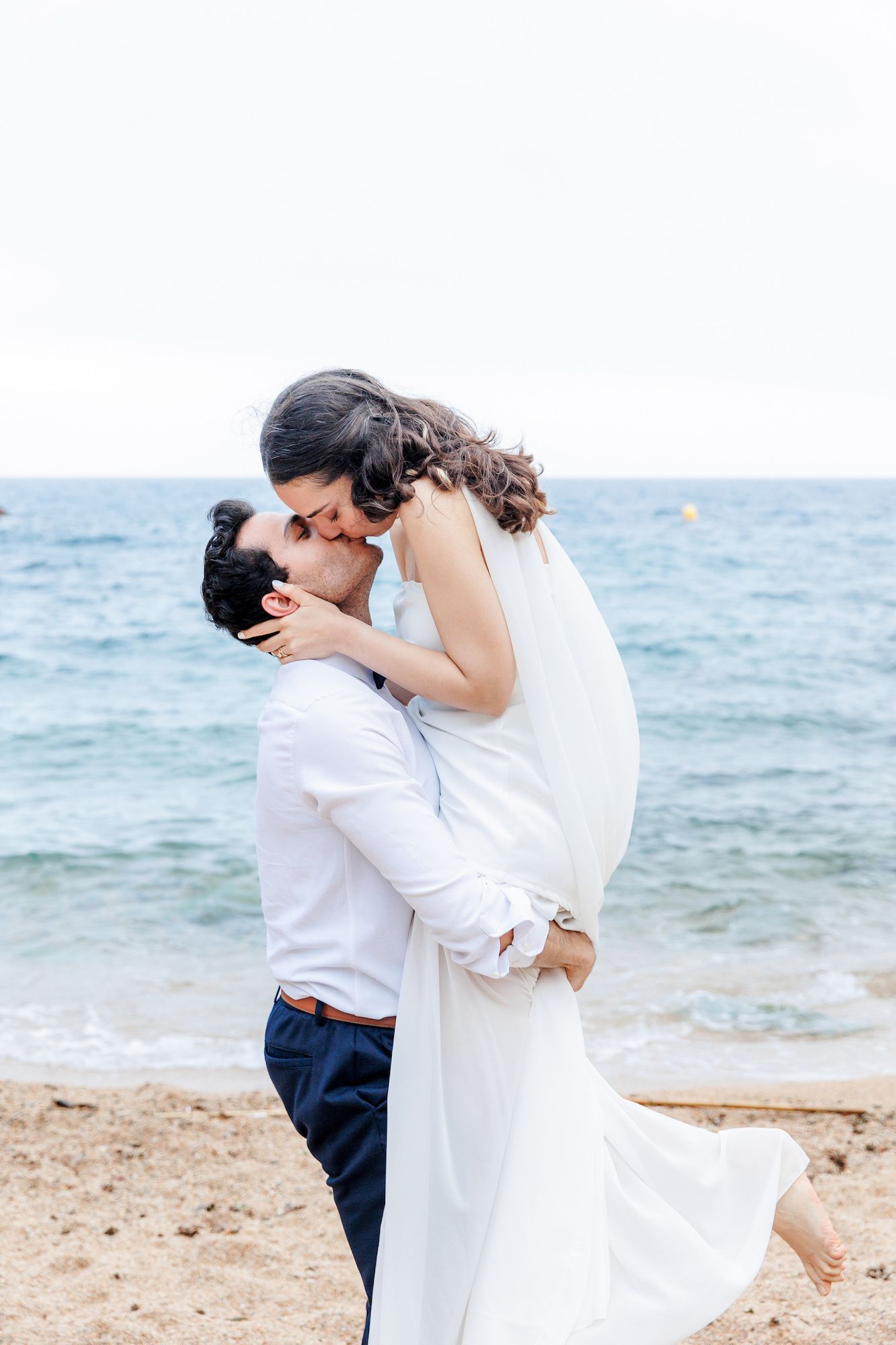 A romantic couple in white attire embraces and kisses on a scenic beach with ocean waves in the background.