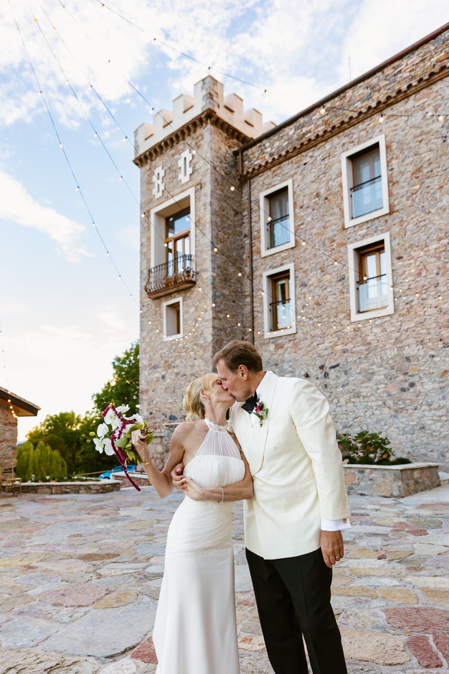 Bride in a white gown holding a bouquet kisses a groom in a white dinner jacket on a stone patio before a castle-style venue.