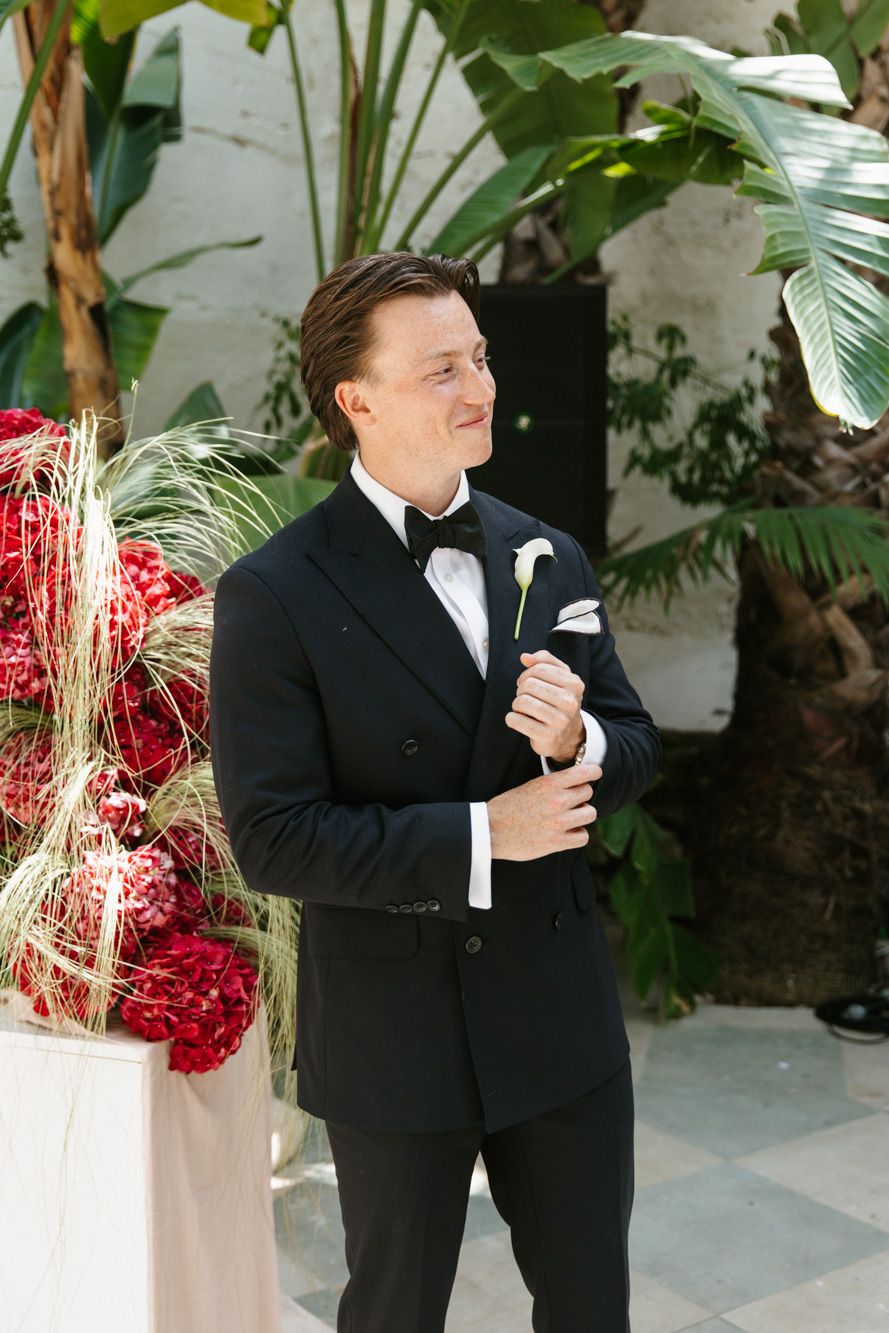 Groom in black tuxedo with white boutonniere smiling outdoors near tropical plants and red floral arrangements.