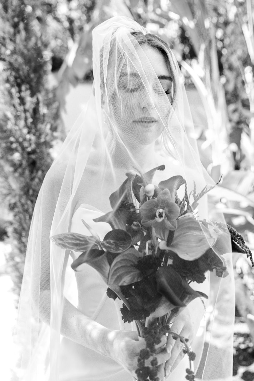 Black and white portrait of veiled bride with eyes closed holding dark floral bouquet outdoors.