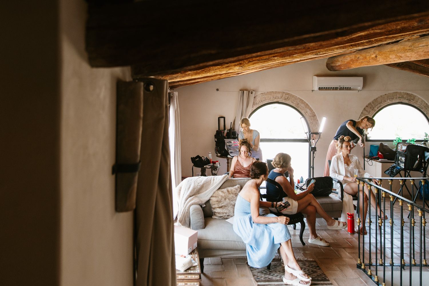 Bridesmaids getting ready in a rustic villa room with arched windows, sitting on a sofa during pre-wedding preparations.
