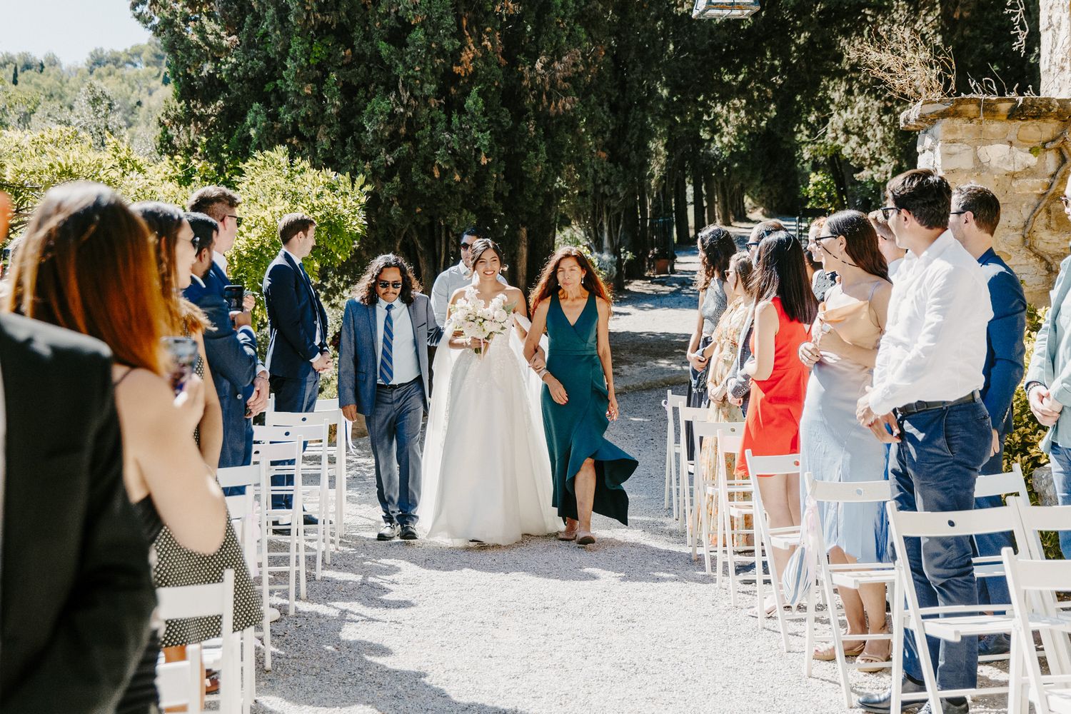 Bride walks down outdoor wedding aisle flanked by guests in white chairs, surrounded by tall trees on a sunny day.