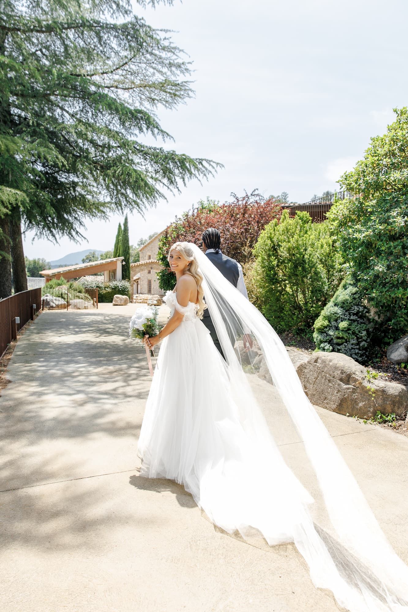 The bride, in a flowing white gown and veil, walks with the groom along a sunny outdoor path at Mas Huix wedding venue
