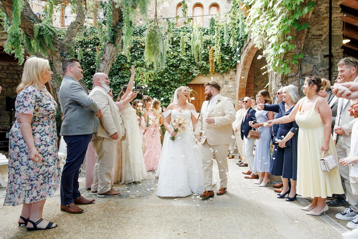 Bride and groom walk through cheering wedding guests in a rustic stone courtyard adorned with lush greenery.